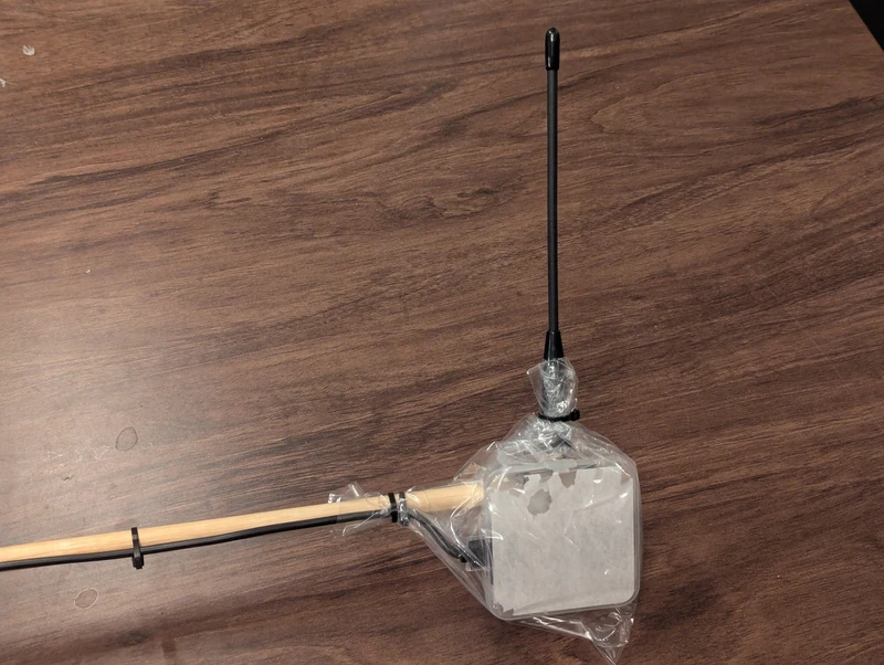 A plastic box encloses a circuit board, attached to a wooden post. A USB cable exits the box and an antenna protrudes out the top. The entire thing is wrapped in clear plastic.