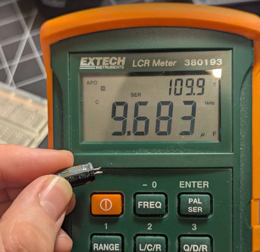 A capacitor labelled 470uF is held next to the display of an LCR meter which reads 9.683uF and 109.9 ohms when measured in series mode at 1kHz.