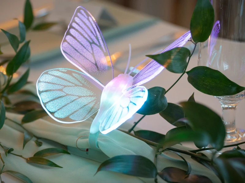The butterfly with glowing acrylic wings situated on a sage green tabletop surrounded by greenery and a wine glass.